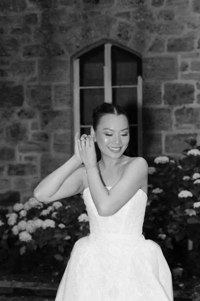 Bridal portrait of a bride putting on earrings by a stone wall and arched window, in a strapless lace ballgown with a delicate bracelet