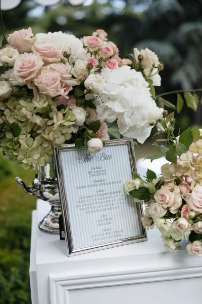 Wedding bar sign in a silver frame listing the cocktail menu, set amid garden trees and a sunlit lawn in an outdoor garden setting.