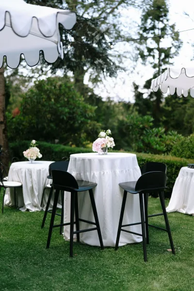 Cocktail hour setup for an outdoor cocktail hour beneath scalloped umbrellas, arranged on a garden lawn bordered by trees and hedges.