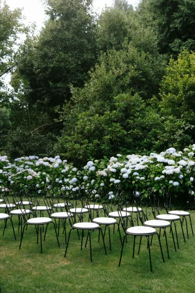 Ceremony seating for a garden ceremony setup in tidy rows of wrought iron chairs on a green lawn, bordered by garden trees and shrubs.