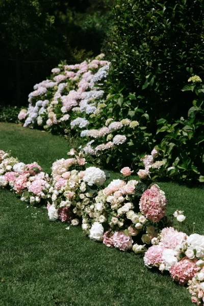 Ceremony aisle flowers create a meandering ground floral aisle of blush hydrangeas, carpeting a grass lawn framed by garden hedges.