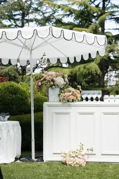 Wedding bar setup shaded by an umbrella for an outdoor wedding bar, framed by garden hedges, trees, and a wide grass lawn under open sky.