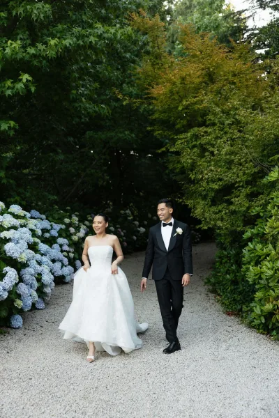 Bride and groom walking in a couple portrait along a gravel garden path bordered by blue hydrangeas, the groom wearing a white rose boutonniere.