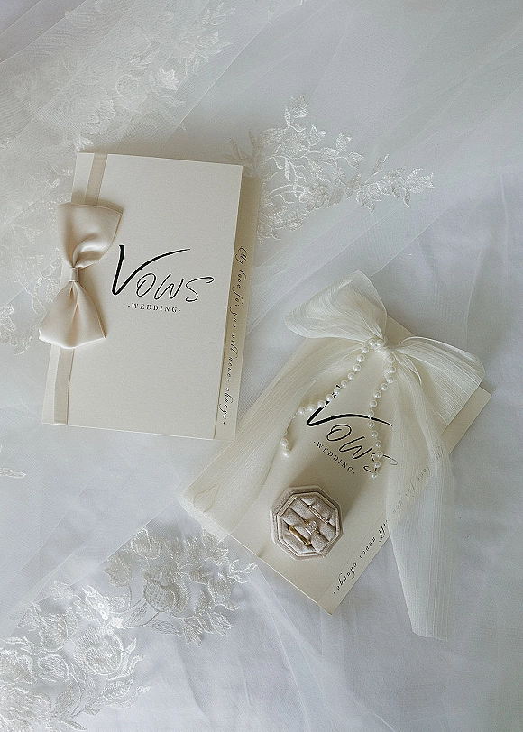 Wedding invitation suite with classic wedding invitations tied in a satin bow, rings and pearl strand on lace fabric over a bridal veil