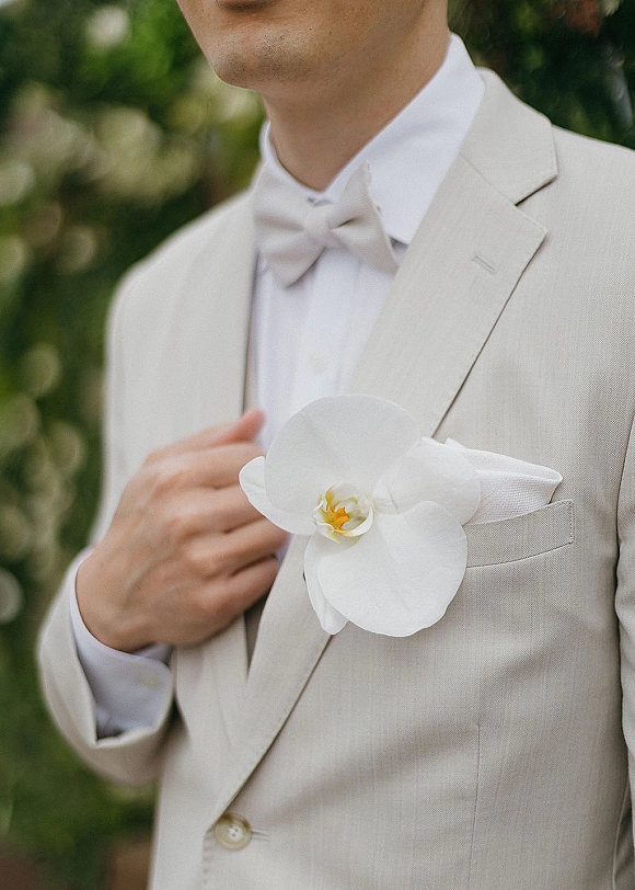 Groom boutonniere featuring a white orchid boutonniere on a beige suit jacket with white bow tie and pocket square, green foliage behind
