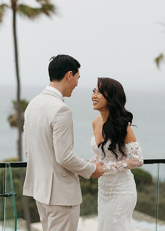 Couple portrait of bride and groom holding hands, bride in lace off-shoulder dress beside groom in suit on oceanfront terrace with palm trees
