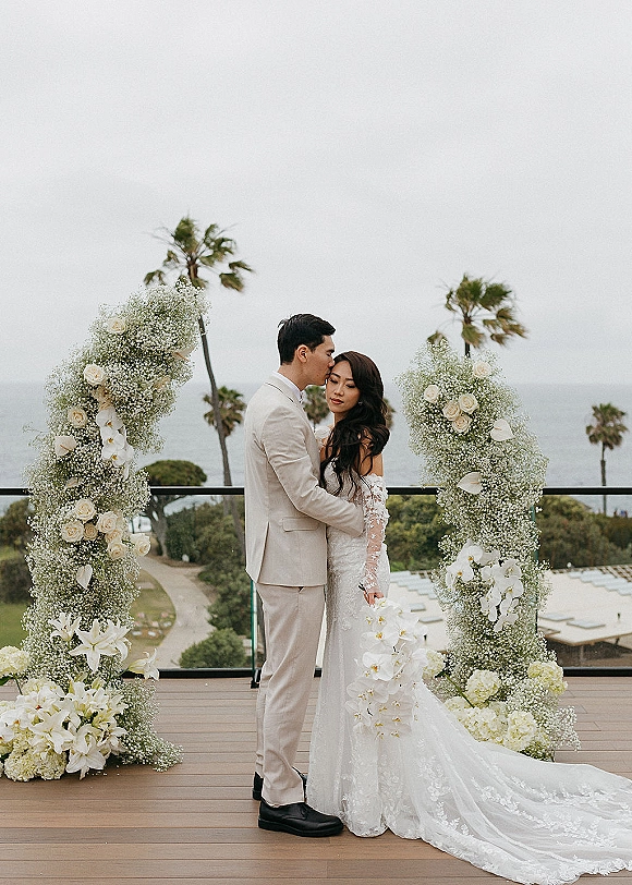 Wedding couple portrait of bride and groom embrace with a forehead kiss beneath a white floral arch on an oceanfront deck with palm trees