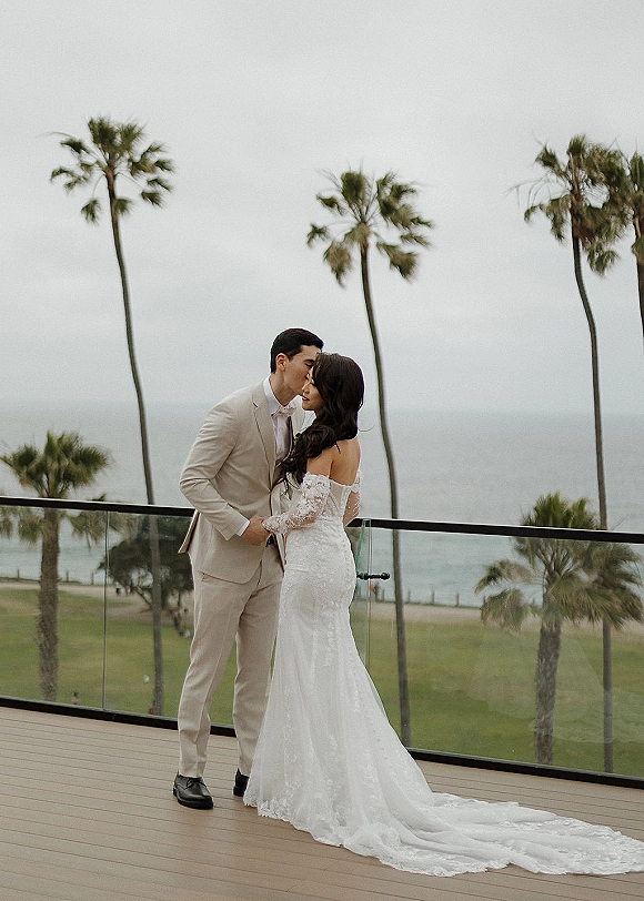 Couple portrait of groom kissing bride’s forehead as she hugs him, lace-sleeve gown with long train on oceanfront terrace with palms