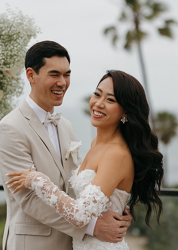 Couple portrait of bride and groom laughing in a newlywed embrace, her strapless lace wedding dress and his beige suit amid blurred greenery