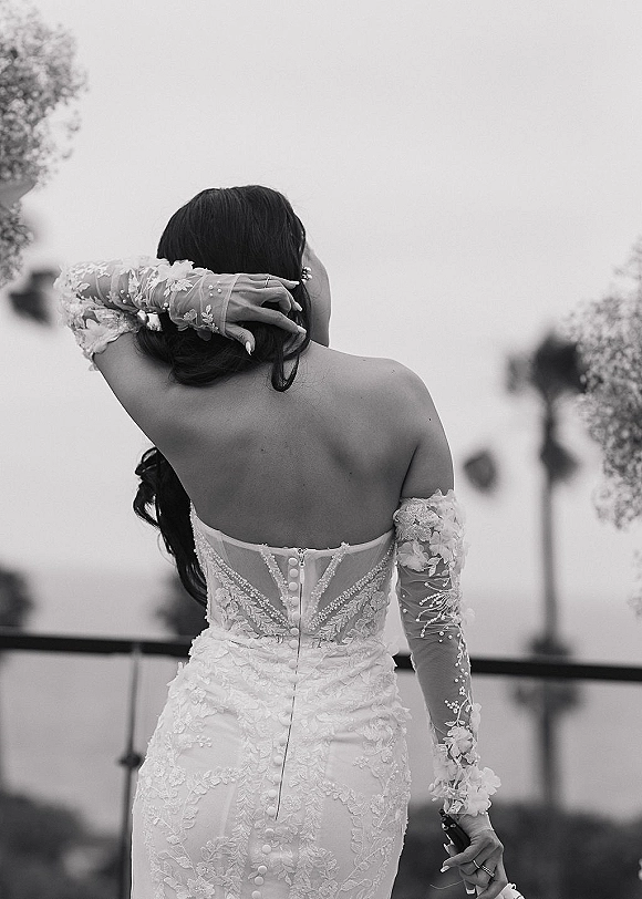 Bridal portrait of a bride from behind adjusting her hair, showing button-back lace off-the-shoulder gown, blurred trees and sky behind