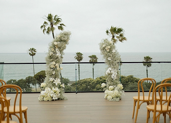 Ceremony setup with white floral columns of orchids and baby's breath, chairs on a wood deck with ocean view and palm trees beyond glass railing