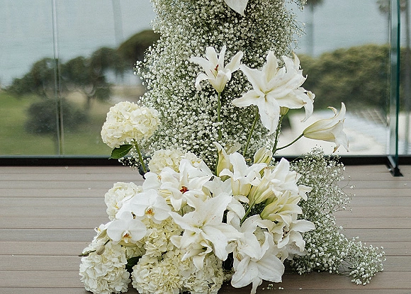 Wedding floral arrangement of white wedding flowers with lilies and hydrangeas on a wooden deck beside glass walls and an outdoor view