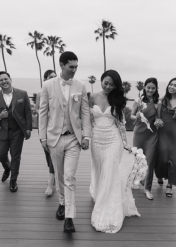 Wedding recessional with bride and groom walking hand in hand on a coastal deck, bride holding a cascading orchid bouquet, ocean and palms behind