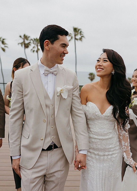 Wedding recessional as bride and groom walking hand in hand, smiling in lace dress and beige suit on a boardwalk with palm trees