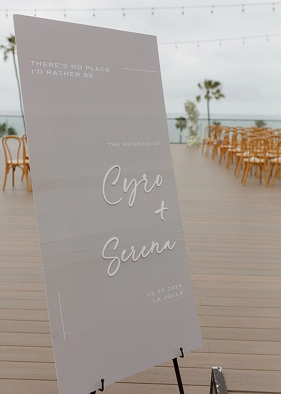 Wedding welcome sign on an easel with modern typography lettering on a wooden deck, with ceremony chairs, string lights, and ocean view