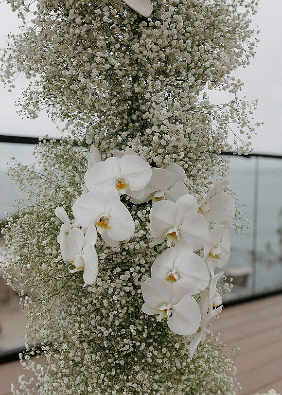 Wedding floral arrangement with baby's breath and cascading white orchids on a patio by a glass railing against the open sky