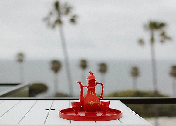 Wedding tea set with a red tea pot and cups on a serving tray atop a patio table, set against an overcast ocean view with palms