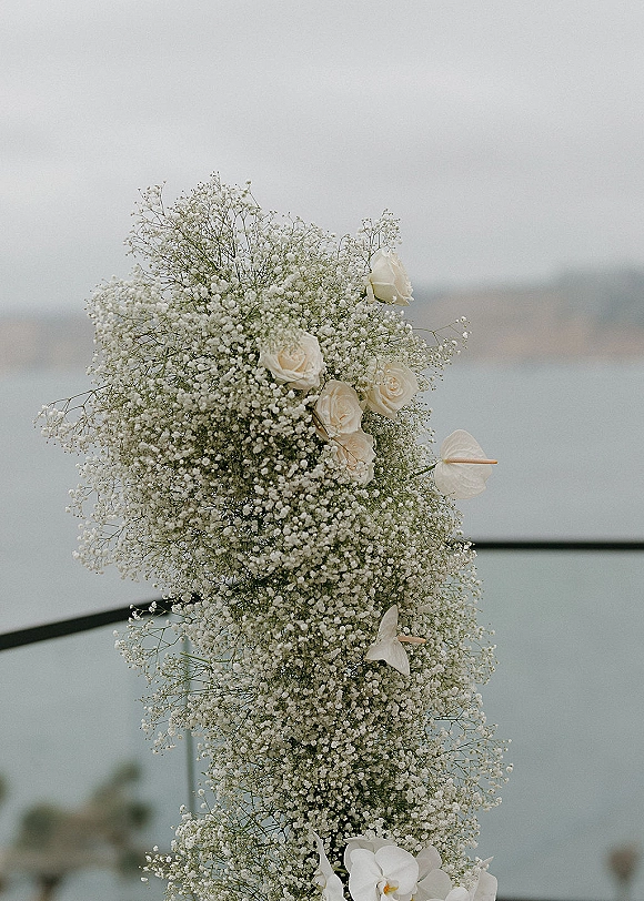 Wedding floral arch with baby's breath and white roses, orchids, and greenery arranged asymmetrically against a cloudy ocean backdrop