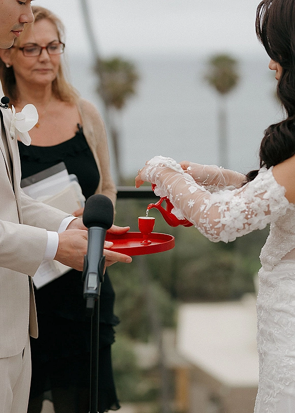 Wedding ceremony ritual with groom holding a red tray and cup as the bride in lace sleeves pours a drink, hills and sky behind