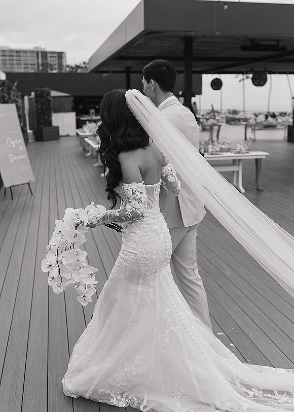 Wedding couple portrait from behind with a long cathedral veil and cascading white orchid bouquet on a rooftop deck with city buildings
