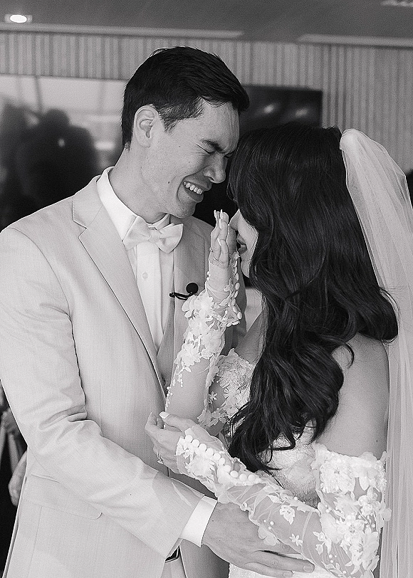 Wedding couple portrait with bride wiping groom tears, her veil framing their faces as he laughs in a white tuxedo by balloons