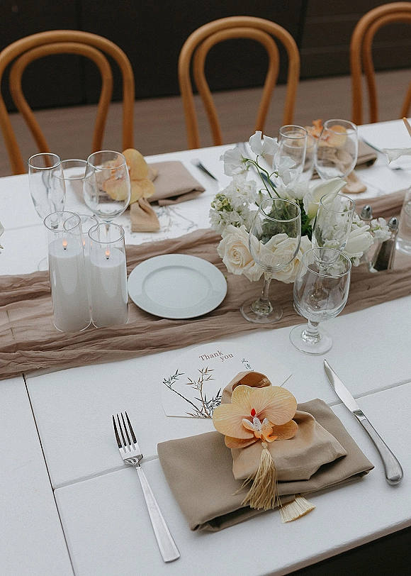 Reception tablescape wedding table setting with white roses and orchids, taper candles in glass cylinders, beige napkins and place cards in an indoor dining space