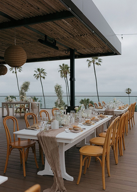 Reception tablescape with a wedding banquet table set with candles and white florals under rattan pendant lights on an oceanfront patio