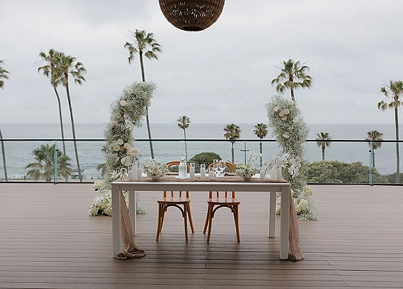Reception sweetheart table with sweetheart table decor under a white floral arch, candles and wooden chairs on an ocean-view terrace with palms