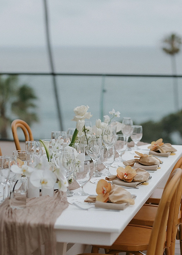 Reception tablescape with a white wedding table, beige gauze runner, orchid place settings, taper candles and bud vases in a glass greenhouse
