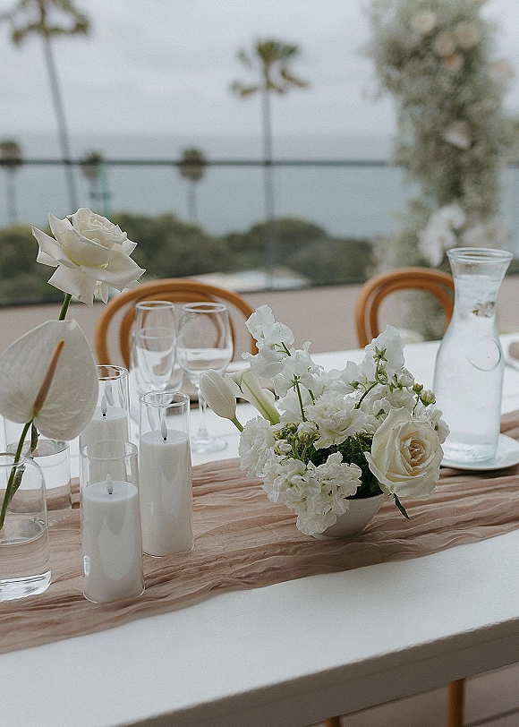 Reception tablescape with a white wedding centerpiece of roses and tulips, taper candles and glassware on a terrace overlooking the ocean