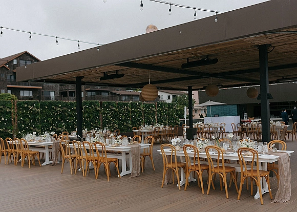 Reception tablescape with long banquet tables wedding, white floral centerpieces, wooden bistro chairs, pendant lanterns and string lights on a covered patio