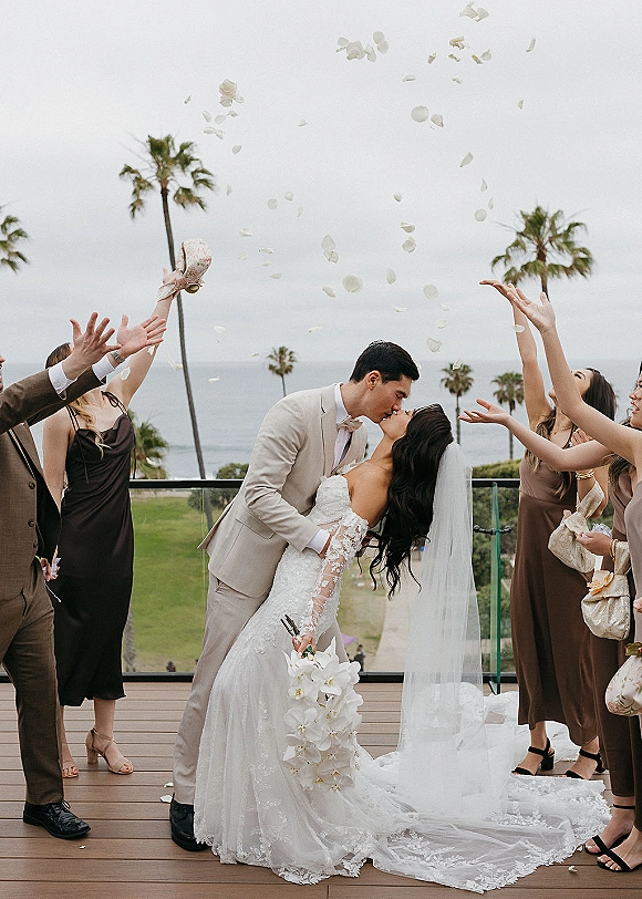Wedding kiss portrait of groom dipping bride in lace dress and veil with bouquet as bridesmaids toss rose petals on an oceanside deck