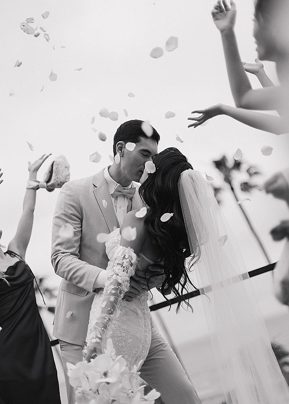 Wedding kiss as guests toss flower petals around the newlyweds, bride in lace dress and veil with palm trees and sky behind