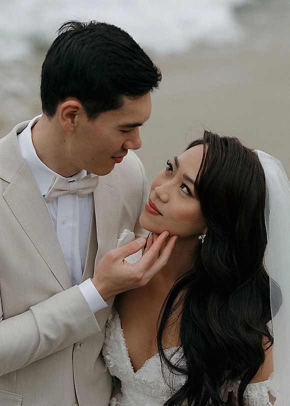 Couple portrait of bride and groom close up as groom touches bride’s face, veil blowing slightly against an outdoor sky backdrop