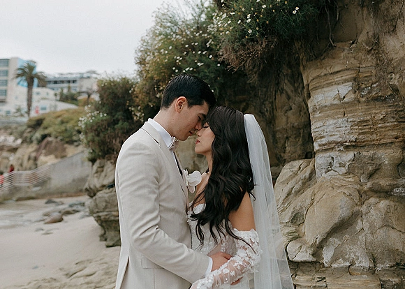 Couple portrait of bride and groom embrace, foreheads touching, her long veil windswept by a rocky beach cliff and ocean shore