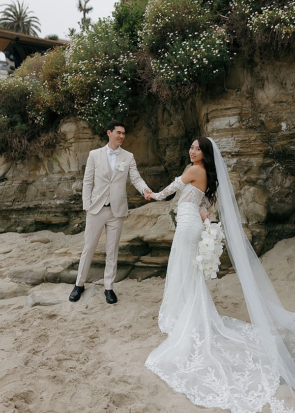 Couple portrait on a sandy beach, bride in a cathedral veil with orchid cascade bouquet, holding hands with groom by rocky cliffs