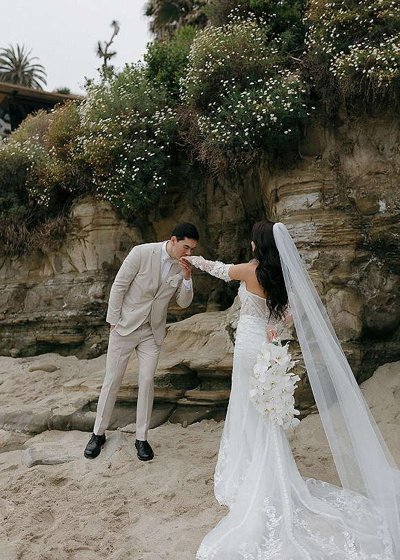 Couple portrait of groom kissing the bride’s hand as she holds an orchid bouquet, long veil flowing on a rocky beach cliffside under overcast sky