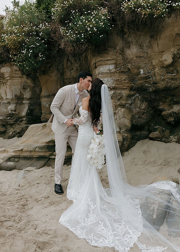 Wedding kiss portrait of bride and groom kissing on a sandy beach, her long veil flowing as she holds a white bouquet by rocky cliffs