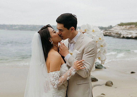 Wedding kiss portrait of bride and groom kissing on a rocky beach, her veil blowing as she holds an orchid bouquet by the ocean