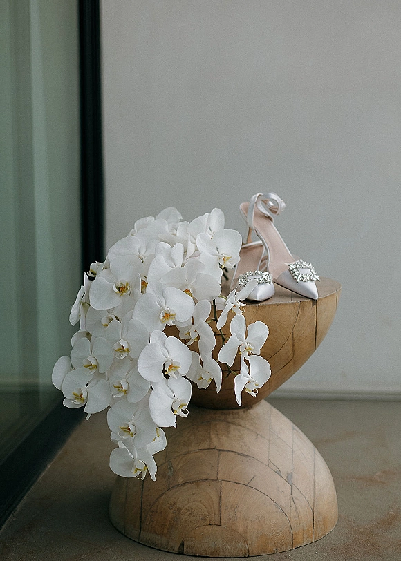Bridal shoes with jeweled buckles and ankle straps on a wooden pedestal, paired with a white orchid accent beside a mirror