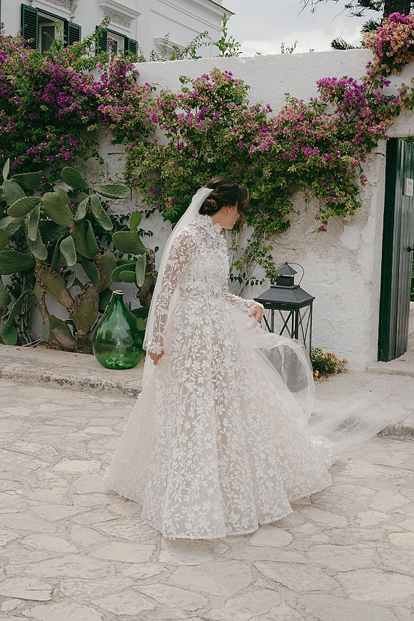 Bridal portrait in a lace wedding dress with a cathedral veil, twirling on a stone patio by a white stucco wall with bougainvillea vines