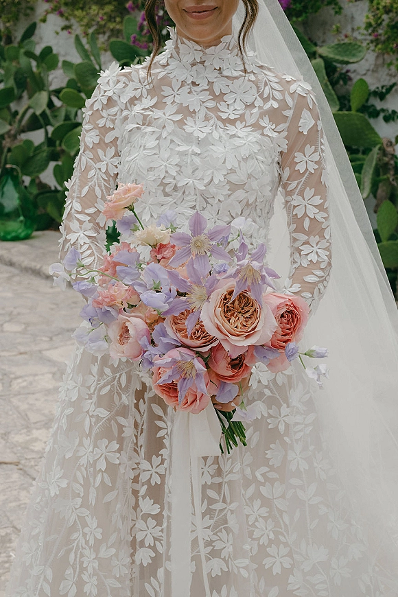 Wedding dress close-up with illusion lace neckline and long sleeves, veil over shoulders, holding a pastel bouquet in a cactus garden backdrop