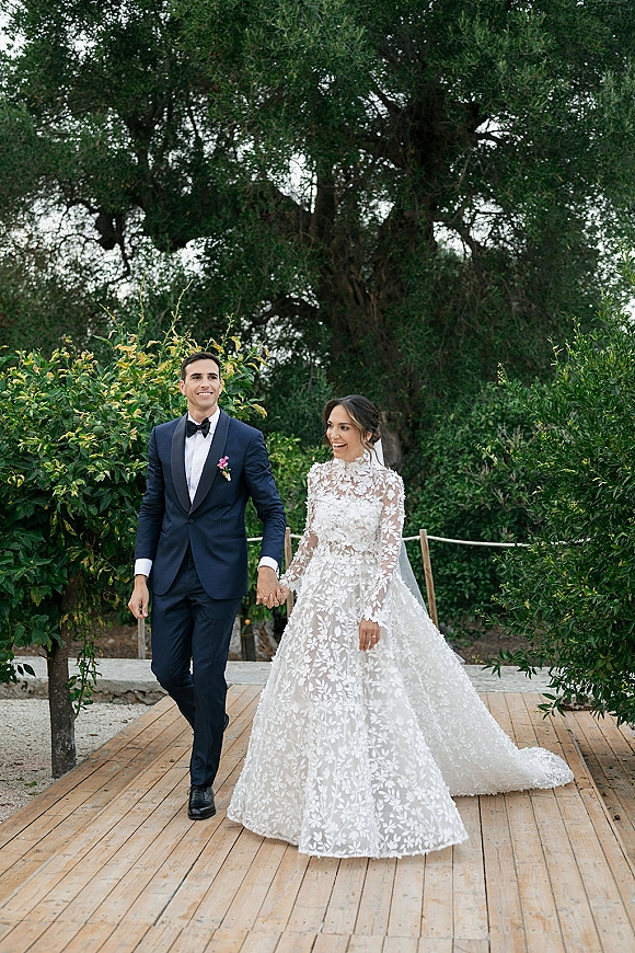 Newlywed couple portrait of bride in long sleeve lace dress and veil holding hands with tuxedoed groom on a garden walkway