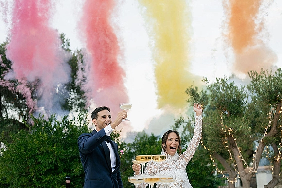 Wedding toast at a champagne tower as bride in lace dress and groom in tux raise coupe glasses under string lights outdoors