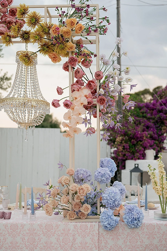 Reception centerpiece with a tall wedding centerpiece floral chandelier of roses, hydrangeas, and orchids above taper candles on a pink patterned tablecloth outdoors