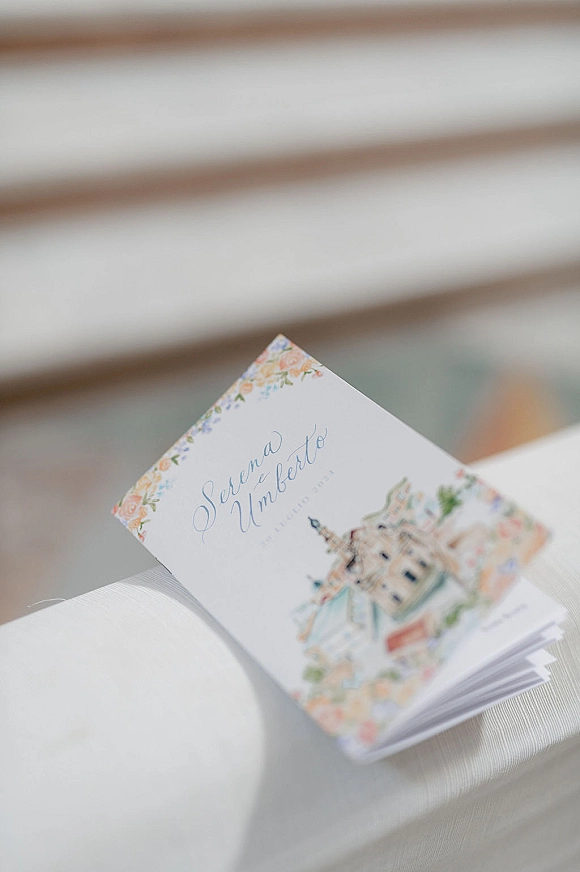 Wedding invitation suite with illustrated invitation card and floral border, paired with white envelopes on a striped chair near neutral siding