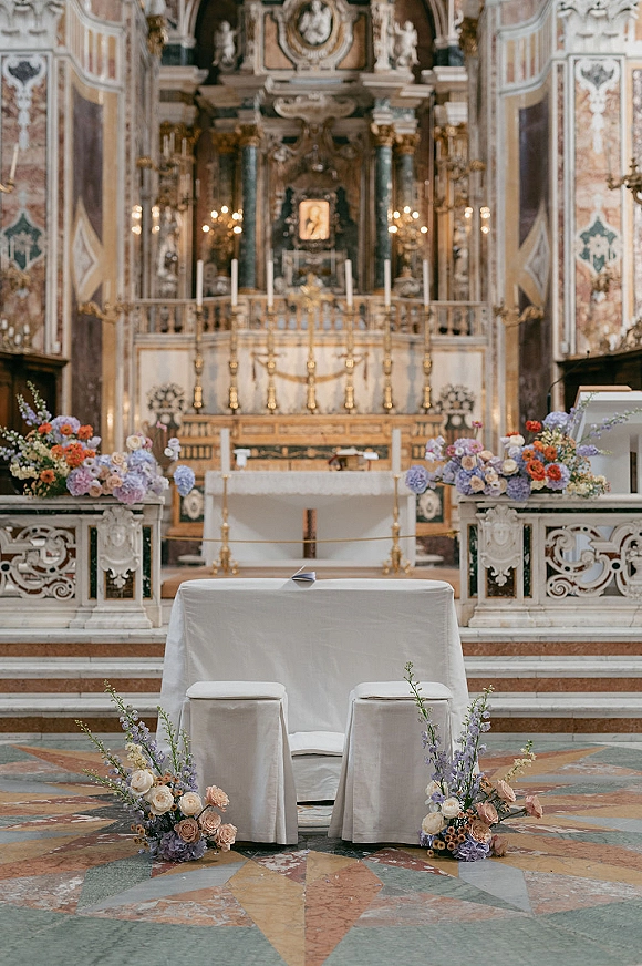 Church ceremony setup with wedding altar flowers on a white-clothed altar table, gold candlesticks, and florals in an ornate marble church interior