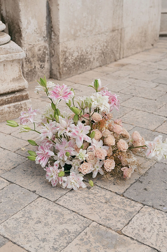 Wedding flowers in a ceremony flower arrangement with pink lilies and blush roses on stone pavement beside a stucco wall and column base