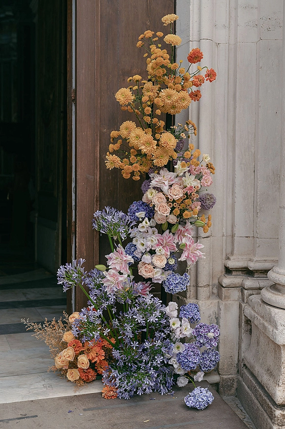 Wedding floral installation with ceremony entrance flowers of roses, lilies, hydrangea and agapanthus cascading on stone steps by a doorway