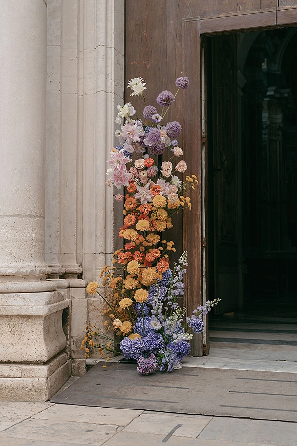 Wedding flower installation with ceremony entrance flowers in pastel, purple and orange blooms with greenery beside a stone doorway and wooden door